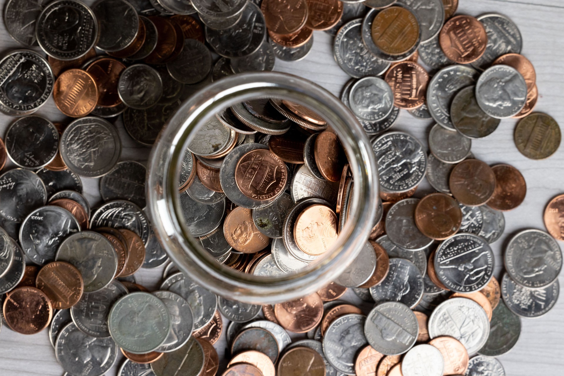 Top view of a jar filled with assorted coins surrounded by scattered change on a wooden surface.