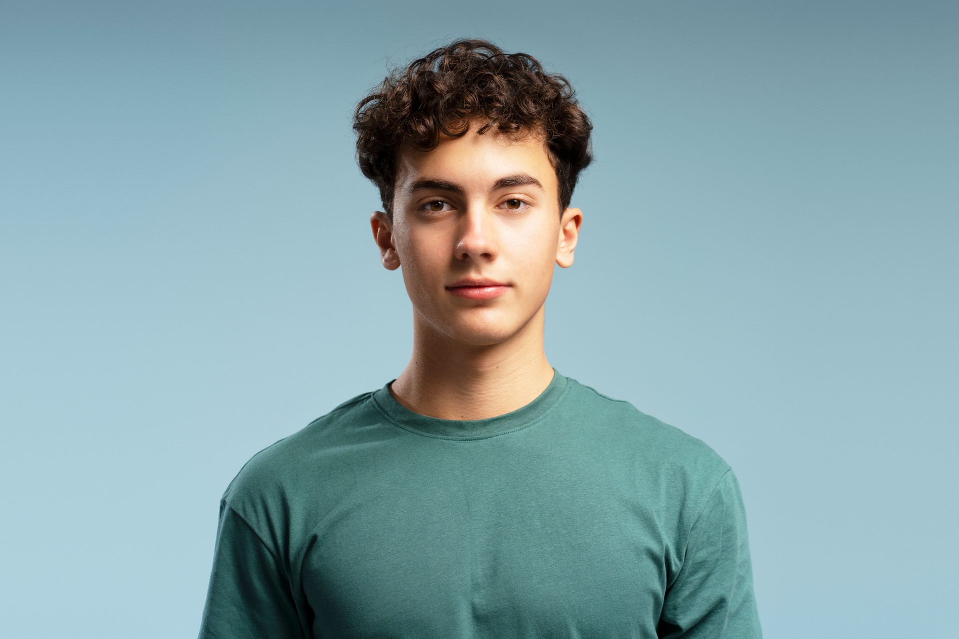 Handsome teen boy with curly hairstyle wearing green t shirt posing isolated on blue background